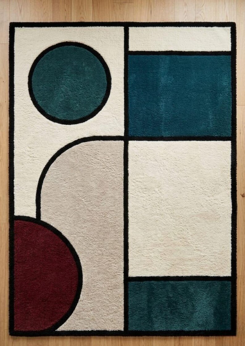 May include: A rectangular area rug featuring a geometric pattern with bold black outlines. The design includes a teal circle, a deep red semicircle, and large blocks of cream, beige, and dark teal. The surface has a soft, textured pile finish.
