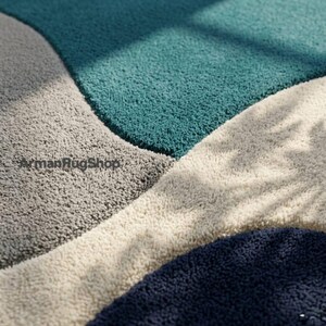 May include: A plush area rug featuring a geometric pattern with large, curved sections in teal, light grey, cream, and dark navy blue. The surface has a thick, shaggy texture. The text ArmanRugShop is printed in black on the grey section.