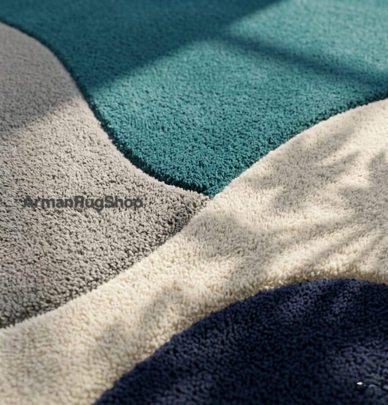 May include: A plush area rug featuring a geometric pattern with large, curved sections in teal, light grey, cream, and dark navy blue. The surface has a thick, shaggy texture. The text ArmanRugShop is printed in black on the grey section.
