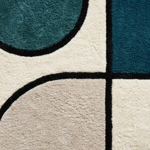 May include: A tufted area rug featuring a geometric pattern of teal, dark blue, and light grey shapes separated by thick black lines on a cream-coloured background. The rug has a dense, textured pile surface with clearly defined colour blocks.