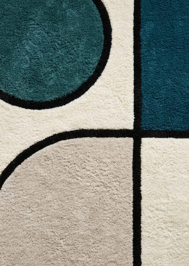 May include: A tufted area rug featuring a geometric pattern of teal, dark blue, and light grey shapes separated by thick black lines on a cream-coloured background. The rug has a dense, textured pile surface with clearly defined colour blocks.