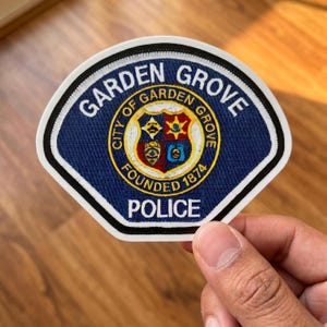 May include: A blue and white Garden Grove Police patch. The patch features the words "Garden Grove" and "Police" in white lettering. The center of the patch has a gold and red emblem with the words "City of Garden Grove" and "Founded 1874".
