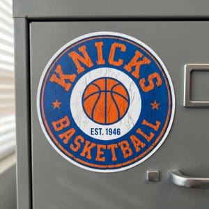 May include: A circular sticker featuring the New York Knicks basketball logo with a distressed texture. The design includes orange text reading KNICKS BASKETBALL, a central orange basketball graphic, and the blue text EST. 1946 on a white background.