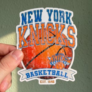 May include: A die-cut sticker featuring a basketball graphic with the text NEW YORK KNICKS in blue and orange. The design includes a smaller team logo, the word BASKETBALL on a banner, and the text EST. 1946, all with a distressed texture.