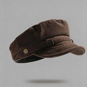 May include: A brown newsboy cap with a structured crown and a short, curved brim. The cap features a decorative strap with a gold-colored button on the side. The hat is made of a soft, textured material.