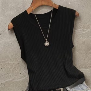 May include: A black sleeveless ribbed top hangs on a wooden hanger. A silver necklace with a pendant is draped over the top. The top is paired with denim jeans.