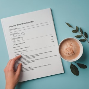 May include: A Cambridge IGCSE Model Paper 2026, with a First Language English section, rests on a light blue surface. A cup of coffee with a foamy top and a sprig of leaves are next to the paper. The paper is being held by a person.