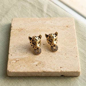 May include: A pair of gold-toned stud earrings shaped like leopard heads. Each earring features a stylized feline face with dark, recessed detailing around the eyes and ears, and a small, circular pendant hanging from the chin.