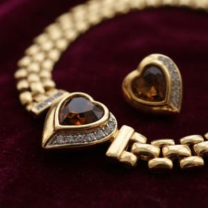 May include: A gold-toned link necklace and a matching single earring, each featuring a heart-shaped brown gemstone surrounded by a border of small clear stones. The necklace chain consists of interconnected rectangular gold-toned metal links.