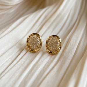 May include: A pair of oval-shaped gold earrings featuring a textured, cross-hatched metallic surface within a smooth, polished gold frame. The earrings have a reflective finish and a distinct, intricate pattern that catches the light.
