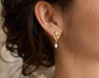May include: A gold-toned earring featuring a tulip-shaped stud with a single white pearl drop dangling from the base. The metal tulip design includes a stem and two leaves, with the pearl suspended from the bottom of the flower by a small link.