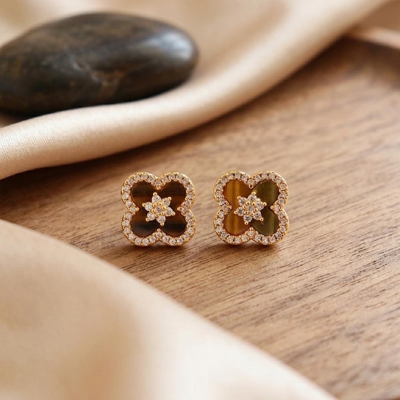 May include: A pair of gold-toned stud earrings shaped like four-leaf clovers. Each earring features a brown tiger's eye stone inlay, a central star-shaped cluster of small clear crystals, and a border of tiny clear crystals around the edge.