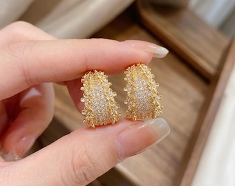 May include: A pair of gold-toned hoop earrings featuring a textured, floral-inspired surface encrusted with numerous small, clear, sparkling stones. The earrings have a wide, curved shape with a dense arrangement of metallic petals and crystals.
