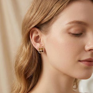 May include: A gold-toned stud earring in the shape of a four-leaf clover. The earring features a brown, smooth center inlay surrounded by a border of small, clear, sparkling stones, with a small star-shaped accent in the very center.
