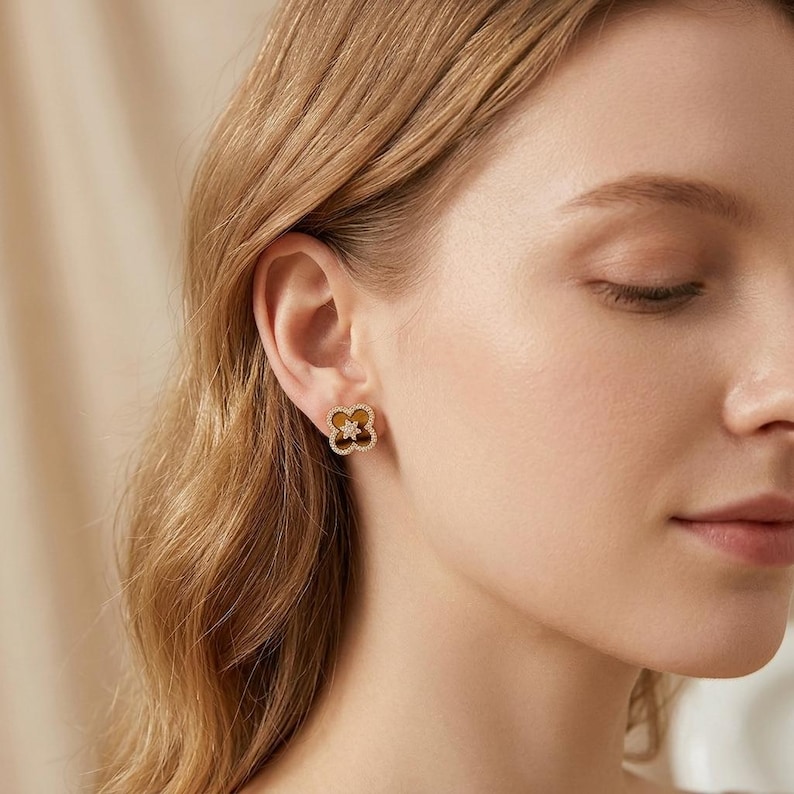 May include: A gold-toned stud earring in the shape of a four-leaf clover. The earring features a brown, smooth center inlay surrounded by a border of small, clear, sparkling stones, with a small star-shaped accent in the very center.