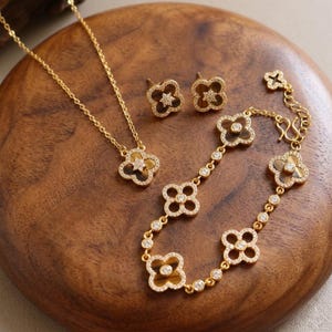 May include: A gold-toned jewellery set consisting of a necklace, a pair of stud earrings, and a bracelet. Each piece features four-leaf clover motifs with brown tiger eye stone centres, surrounded by small clear crystals set in gold-toned metal.