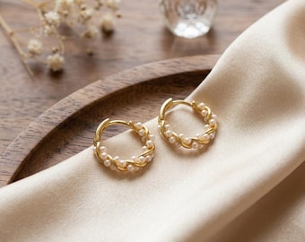 May include: A pair of gold hoop earrings featuring a twisted design adorned with small white pearls. The earrings are circular in shape with a hinged closure mechanism, showcasing a metallic gold finish and a series of evenly spaced round pearls.