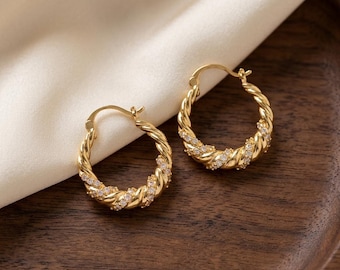 May include: A pair of gold hoop earrings featuring a twisted rope design. Each hoop is accented with small, clear, sparkling stones embedded along the twisted metal surface, creating a textured appearance with a polished metallic finish.