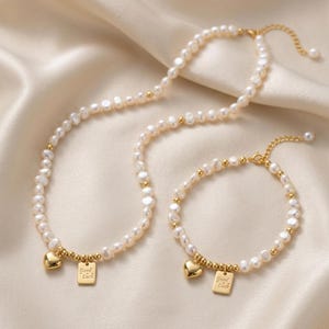 May include: A necklace and bracelet set featuring white freshwater pearls interspersed with small gold-toned beads. Each piece includes a gold-toned heart charm and a rectangular gold-toned pendant engraved with the text Good Luck.