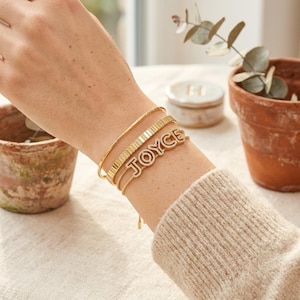 Op de afbeelding: Een set van drie goudkleurige armbanden om de pols. De eerste is een gladde dunne armband, de tweede is een schakelketting met textuur en de derde is een fijne kettingarmband met een goudkleurig naamplaatje met de naam JOYCE in kristallen.