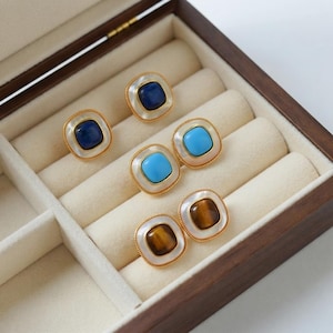 Peut inclure: Trois paires de boucles d'oreilles carrées avec des pierres centrales bleu foncé, turquoise vif et œil-de-tigre marron. Chaque pierre est entourée d'une bordure blanche et d'un fin contour en métal doré, disposées dans un coffret à bijoux en bois.