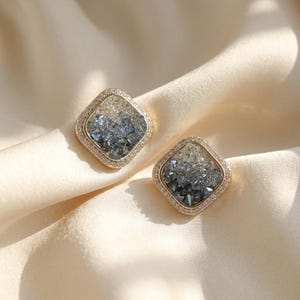 May include: A pair of square-shaped stud earrings featuring a gold-toned metal border encrusted with small clear crystals. The center of each earring contains a gradient of faceted stones transitioning from clear at the top to dark blue at the bottom.