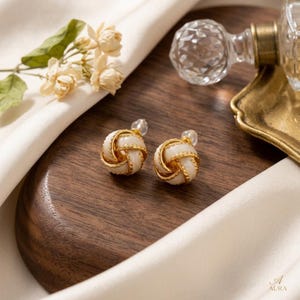 May include: A pair of stud earrings featuring a spherical knot design. Each earring consists of a smooth, cream-colored center intertwined with a textured, gold-toned metal rope detail. The earrings have clear plastic backings for secure attachment.