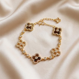 May include: A gold-tone chain bracelet featuring four clover-shaped charms with brown centers and clear crystal accents. The bracelet includes small circular links set with clear crystals between each clover charm and an adjustable chain extension.