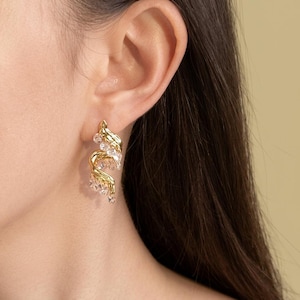Könnte beinhalten: Ein goldfarbener Ohrring mit einer geschwungenen, organischen Metallstruktur, die mit kleinen, klaren, facettierten Kristallen besetzt ist. Das Design hat eine vertikale, kaskadierende Form, die sich dem Ohrläppchen anpasst und eine polierte Oberfläche aufweist.