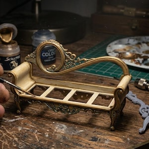 Può includere: Una chaise longue in miniatura nei toni crema e oro. La chaise longue presenta dettagli di volute e viene dipinta con un pennello sottile. Vernici e strumenti sono visibili sulla superficie di lavoro in legno.