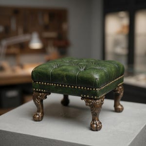 May include: A green leather ottoman with button tufting and brass nailhead trim. The ottoman has ornate, bronze-colored legs with a claw foot design. The background is blurred, suggesting a workshop or studio setting.