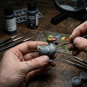 May include: A miniature gray duck figurine being hand-painted with a fine brush. The duck has a yellow and green head, a red beak, and yellow spots on its body. Paint bottles, brushes, and a magnifying glass are in the background.