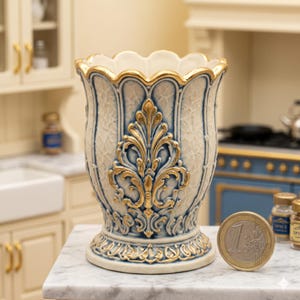 Könnte beinhalten: Eine verzierte, cremefarbene Vase mit goldenen und blauen Akzenten. Die Vase hat ein detailliertes, erhabenes Blumendesign und einen gewellten Rand. Die Vase ist etwa 10 cm hoch und steht auf einer Marmoroberfläche.