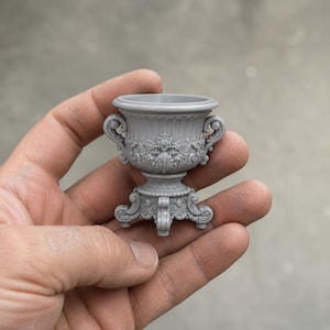 May include: A miniature gray urn-shaped planter featuring intricate relief carvings of floral motifs and scrollwork. The vessel has a wide circular rim, two side handles, and a decorative pedestal base with four small feet, all in a matte finish.
