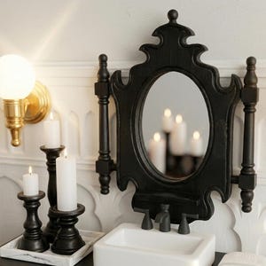 Puede incluir: Un espejo negro con marco ornamentado y espejo ovalado. El espejo está montado sobre un lavabo blanco con grifos negros. Tres candelabros negros con velas blancas están sobre una bandeja. Un aplique dorado con una bombilla redonda está en la pared.