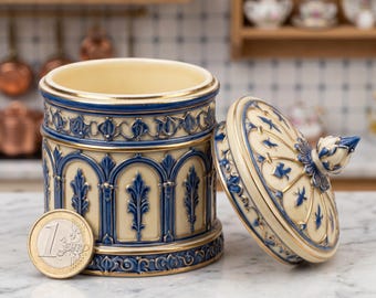 Può includere: Un vaso decorativo in ceramica con coperchio, con base color crema e dettagli blu e oro. Il design del vaso include motivi ad arco e floreali. Una moneta da euro è accanto al vaso per il confronto delle dimensioni. Altezza circa 10 cm.