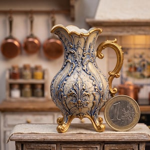 May include: A decorative, ornate pitcher with a gold handle and base, featuring a cream and blue floral design. The pitcher is displayed on a wooden surface, with a 1 Euro coin next to it for scale. The background includes kitchen elements.
