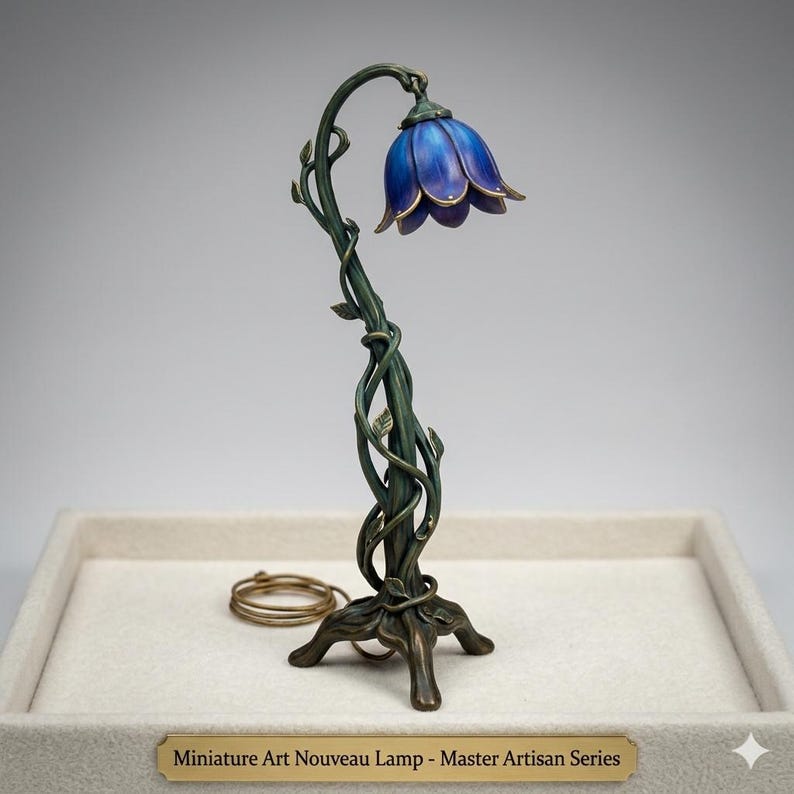 Może przedstawiać: Miniaturowa lampa w stylu Art Nouveau z niebieskim kloszem w kształcie kwiatu i brązową podstawą. Trzon lampy przypomina winorośl z liśćmi. Na podstawie widnieje napis: "Miniature Art Nouveau Lamp - Master Artisan Series."