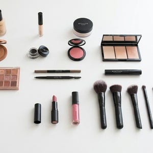 Puede incluir: Una variedad de productos de maquillaje, incluyendo base, corrector, bronceador, paletas de sombras de ojos, colorete, delineador de ojos, lápices de cejas, lápiz labial, brillo labial y brochas de maquillaje, dispuestos sobre una superficie blanca.