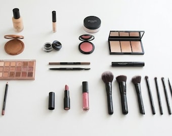 Può includere: Un assortimento di prodotti per il trucco, tra cui fondotinta, correttore, bronzer, palette di ombretti, fard, eyeliner, matite per sopracciglia, rossetto, lucidalabbra e pennelli per il trucco, disposti su una superficie bianca.