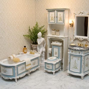 May include: A miniature bathroom suite featuring a bathtub, toilet, vanity unit, and cabinet. The furniture is white with pale blue detailing and gold trim. Floral patterns decorate the doors and panels. A small statue and potted plant complete the scene.