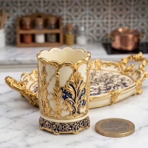 Könnte beinhalten: Eine verzierte, cremefarbene Tasse mit goldenen und blauen Akzenten, mit einem gewellten Rand und aufwendigen floralen Designs. Ein passendes Tablett mit ähnlichen Details befindet sich hinter der Tasse. Eine Euro-Münze liegt neben der Tasse.