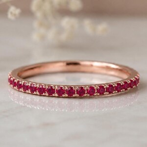 May include: A thin rose gold band ring featuring a continuous row of small, round, vibrant red rubies set along the outer circumference. The metal has a polished finish, and the stones are uniform in size and colour, creating a circular design.