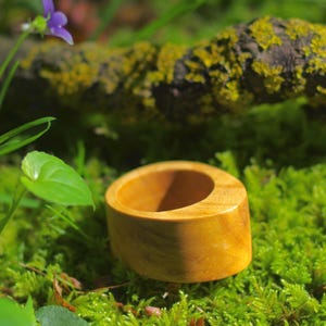 May include: A smooth, light brown wooden ring with a slanted, asymmetrical top edge. The ring features a polished finish that highlights the natural grain patterns of the wood, showcasing a circular opening and a thick, solid band construction.