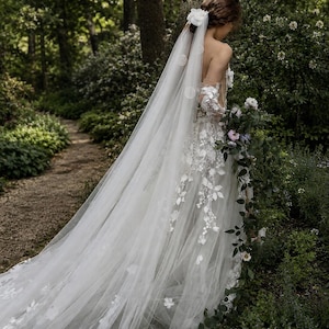 May include: A floor-length white wedding dress featuring intricate floral lace appliqués, a strapless bodice, and long sheer sleeves. A long, flowing tulle veil is attached to the hair with a white floral accessory, cascading down the back.