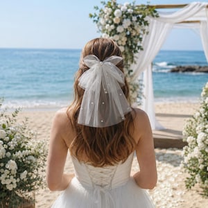 May include: A white tulle hair bow veil featuring scattered small white pearls. The accessory is styled as a bow with a short, layered veil extending downwards, attached to a hair clip, designed to be worn in the hair for a decorative look.