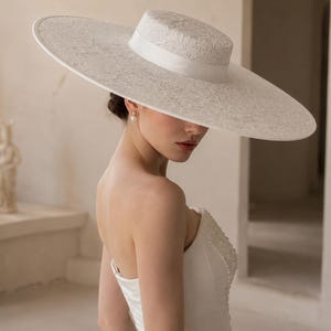 May include: A wide-brimmed ivory hat featuring a flat crown and a delicate lace overlay. The hat is accented with a smooth, solid-colored ribbon band encircling the base of the crown, providing a clean and structured appearance to the headwear.