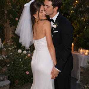 Puede incluir: Un vestido de novia blanco sin tirantes con una hilera de botones en la espalda, combinado con un velo de tul blanco transparente con un lazo en la parte superior. El conjunto se lleva con un esmoquin negro con un prendido blanco y pajarita.