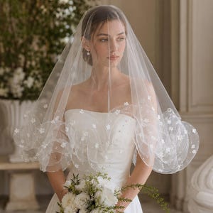 May include: A sheer white bridal veil featuring a ruffled edge adorned with small white floral appliqués and scattered clear beads. The veil is draped over the head and shoulders, showcasing a delicate layered design with a soft, translucent finish.