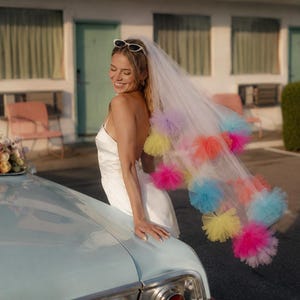 Könnte beinhalten: Ein weißer Brautschleier mit verstreuten, bunten Tüll-Pompons in den Farben leuchtendes Pink, Orange, Gelb und Hellblau. Der Schleier besteht aus einem transparenten, leichten Netzstoff und ist als dekoratives Accessoire für den Kopf konzipiert.
